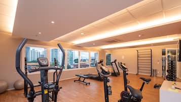 Un gimnasio con equipo de ejercicio diverso, amplias ventanas y vista a edificios.
