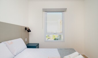 Una habitación de hotel con cama, mesita de noche, lámpara y ventana con vistas a la ciudad.