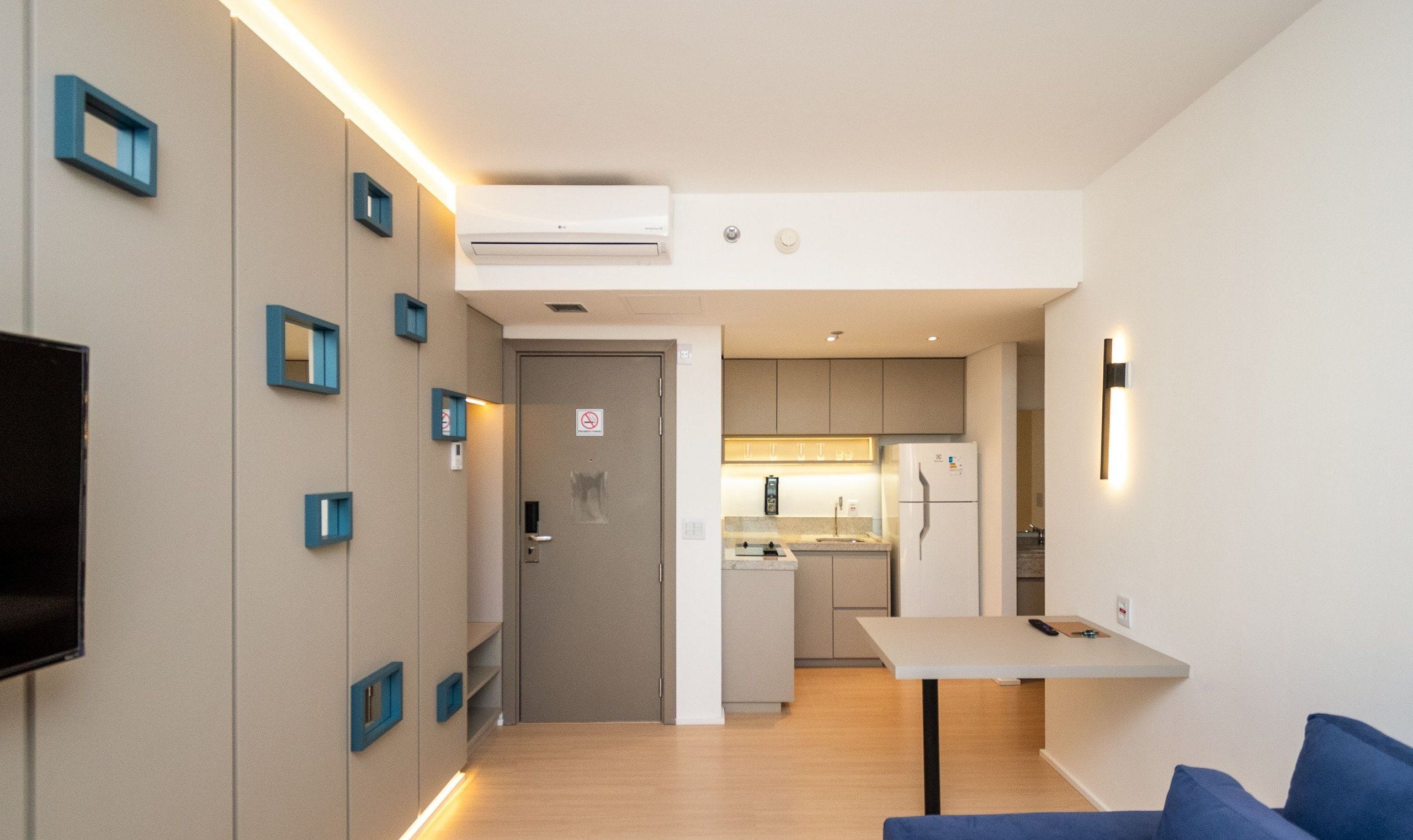 A compact hotel room with a kitchenette, a flat-screen TV mounted on the wall, and a small seating area with a table and blue cushions.