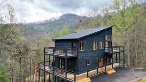 A two-story cabin with a balcony, surrounded by forested hills.