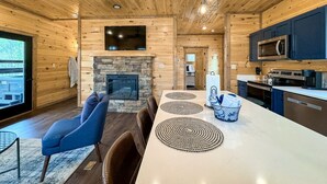 A modern log cabin interior with a stone fireplace, a dining area with placemats, a kitchen with stainless steel appliances, and a large TV mounted above the fireplace.