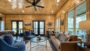 A cozy cabin interior with a wooden ceiling, a ceiling fan, a sofa, a coffee table, and a dining area.