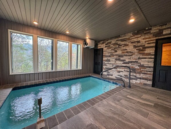 An indoor swimming pool with a stone wall and wooden ceiling, large windows, and a door. - New! Modern Pool Cabin in Sevierville Cabin (Sevierville)