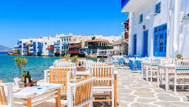 A seaside restaurant with outdoor seating, white and blue buildings, and a view of the sea.