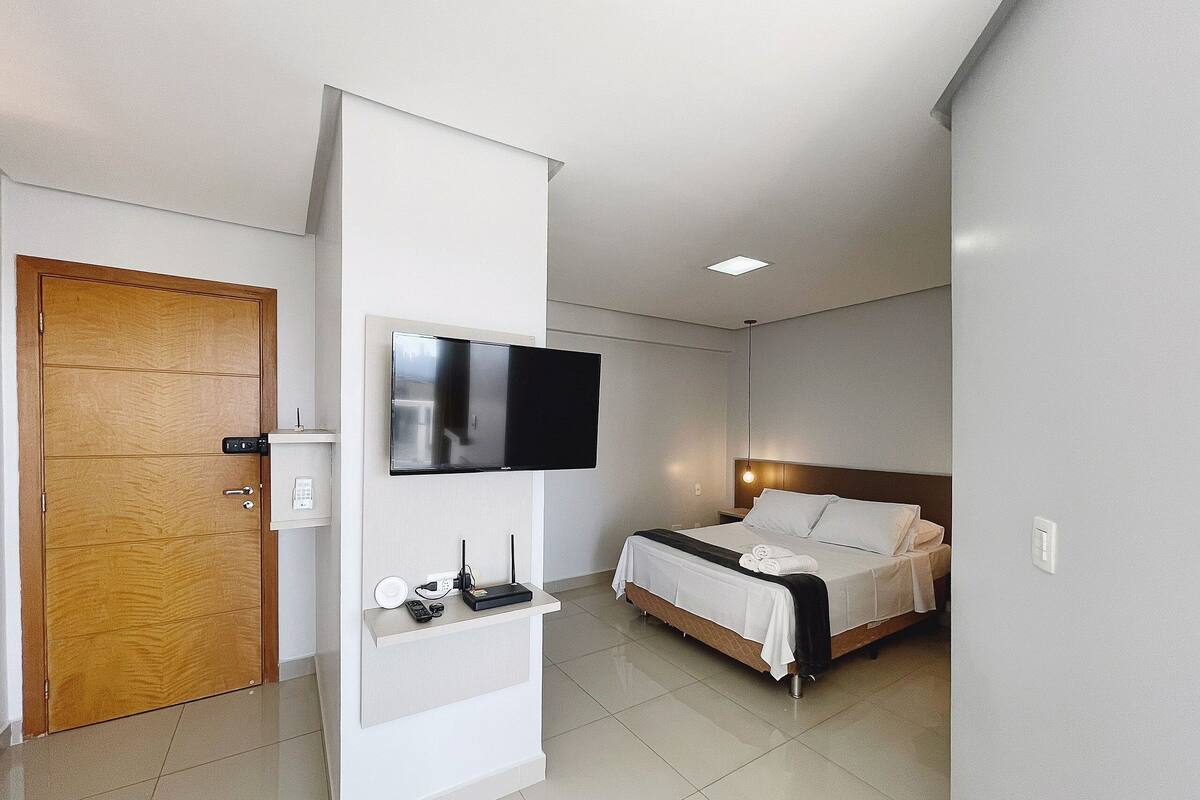 A modern hotel room with a wooden door, a flat-screen TV mounted on a white wall, a bedside table with a remote control, and a neatly made bed with white linens.