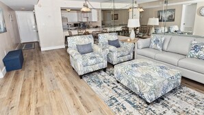 A modern living room with a sofa, two armchairs, a coffee table, and a rug. There is a kitchen in the background with cabinets and a dining area.