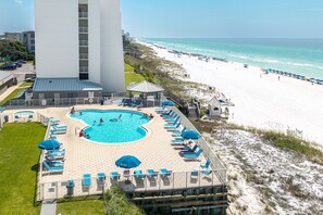 A pool area with sun loungers and umbrellas, a beach with umbrellas and people, and a tall building in the background. - BH 402D Beautiful Views from this 2 bed/2 bath (Miramar Beach)