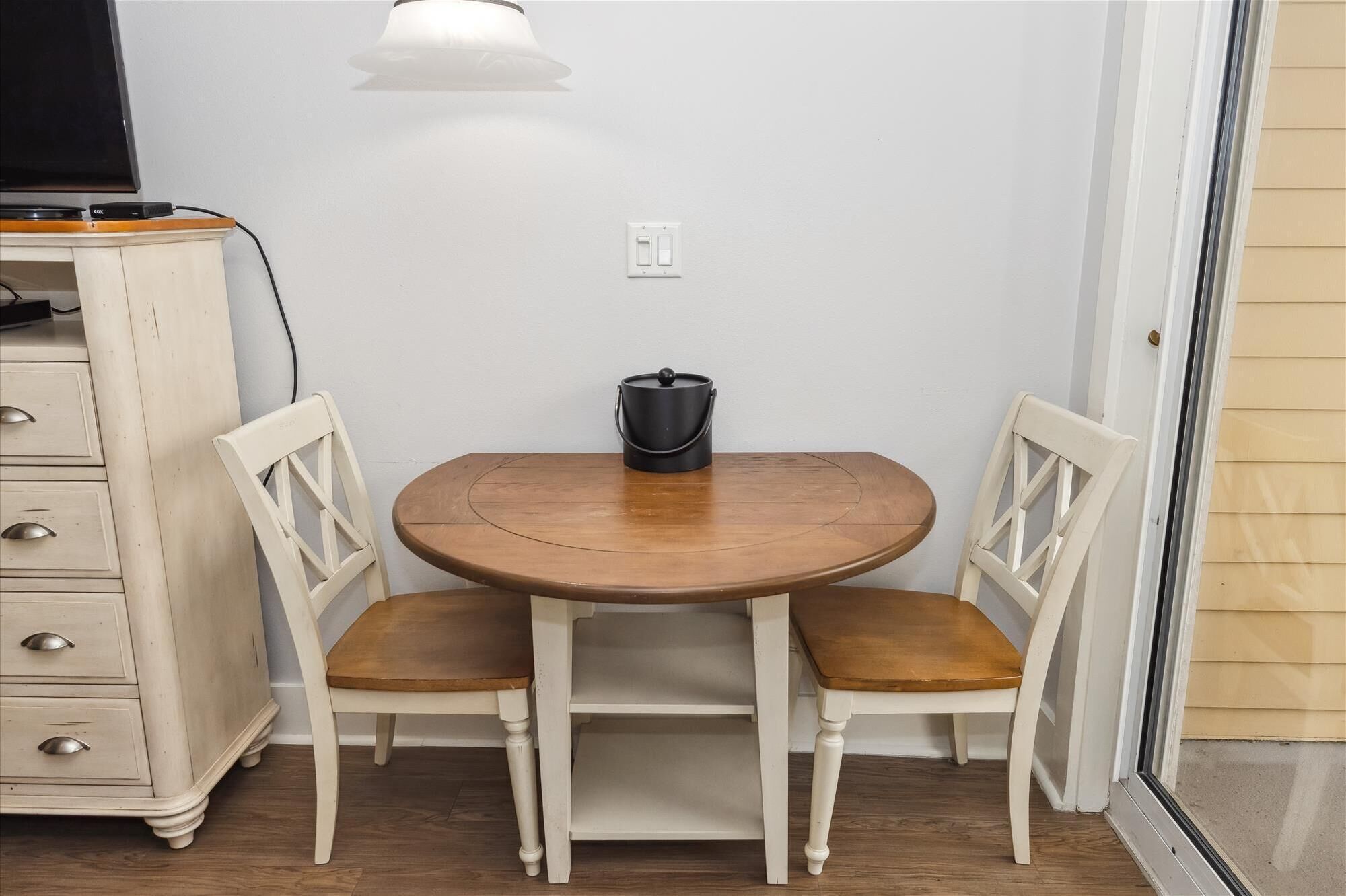 A small dining area with a round wooden table and two white chairs. There is a black pot on the table, a white wall, and a wooden cabinet with drawers to the left.