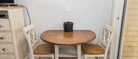 A small dining area with a round wooden table and two white chairs. There is a black pot on the table, a white wall, and a wooden cabinet with drawers to the left.