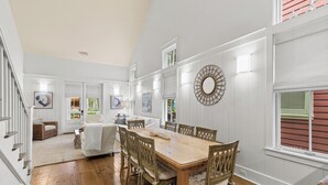 Uma sala de jantar ampla, com uma mesa e cadeiras de madeira, cercada por painéis brancos e grandes janelas.