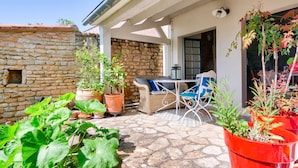 A stone wall, a wicker sofa, and a table with chairs in a covered outdoor area. - Loix- Ré Island- House for 5 (Loix)