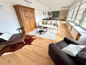 A living room with a sofa, a leather chair, a wooden cabinet, a dining area with a table and chairs, and a kitchen in the background.