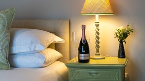 Una cama bien tendida con sábanas blancas, una botella de Bollinger, una copa, una lámpara y un florero con flores sobre una mesita de noche.