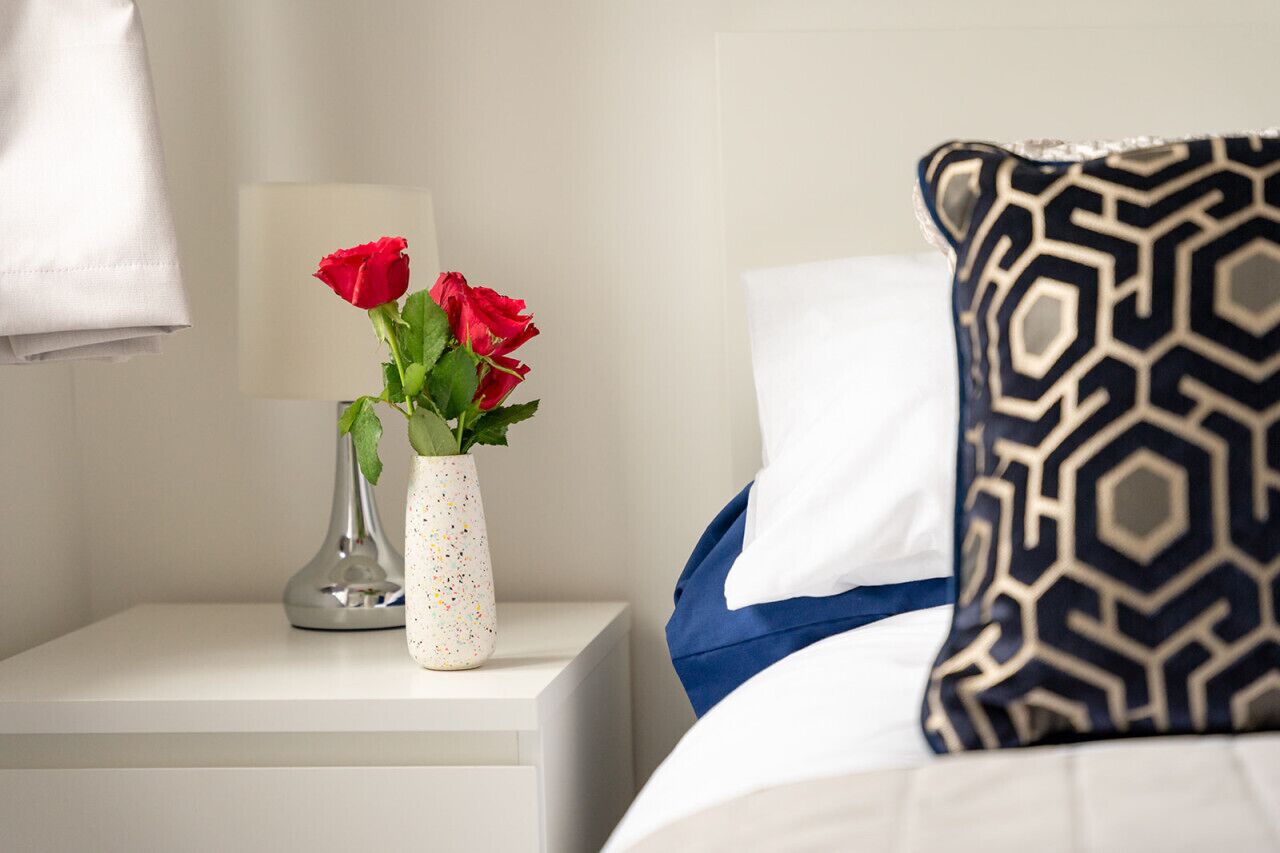 Un lit bien fait, agrémenté d’un coussin à motifs, une table de chevet blanche avec un vase de roses rouges et une lampe.