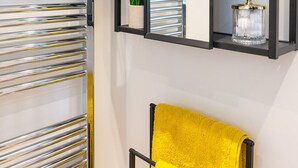 A modern bathroom with a chrome towel rack, a yellow towel, and a black-framed mirror with shelves holding a plant and a glass jar.