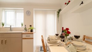 A modern kitchen with white cabinetry, a dining area with a table set for two, and a window with blinds.