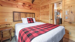 A wooden cabin room with a bed, a small wooden table, and a kitchen area visible through an open door. - Grassy Cove- Cozy Smoky Mtn Cottage w/Hot Tub (Sevierville)