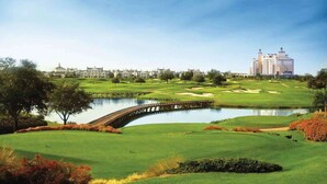 A golf course with a bridge over a pond, surrounded by lush greenery and a large hotel in the background.