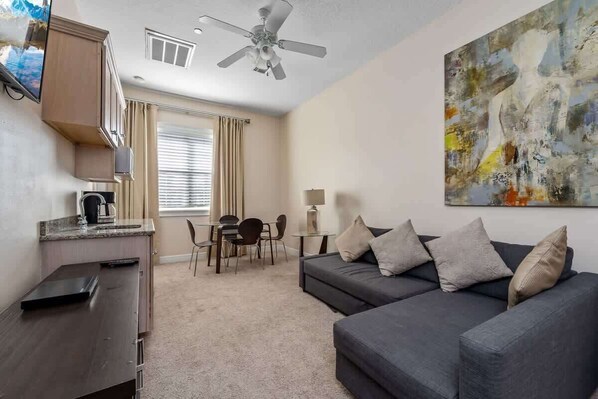 A modern living room with a dark gray sofa, a dining table with chairs, a ceiling fan, and a large abstract painting on the wall.