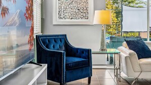 A modern living room with a blue velvet armchair, a white sofa, a glass coffee table, and a framed abstract art piece. - 9BR villa w/ pool, hot tub & patio (Kissimmee)
