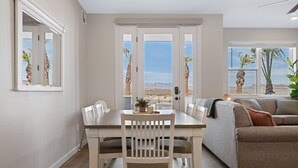Een zithoek met een tafel en stoelen, een bank, en uitzicht op palmbomen en bergen door grote ramen.