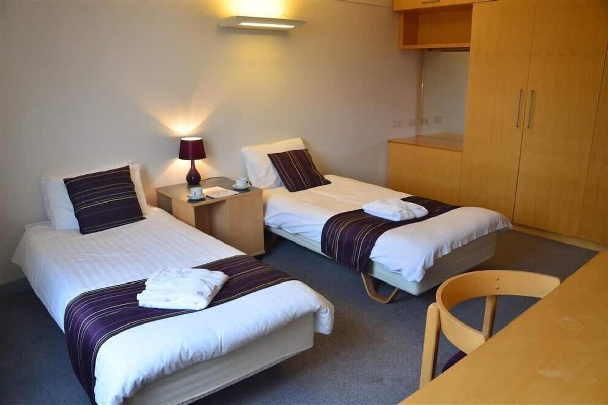 A hotel room with two single beds, a wooden desk, a chair, and a small kitchenette area.