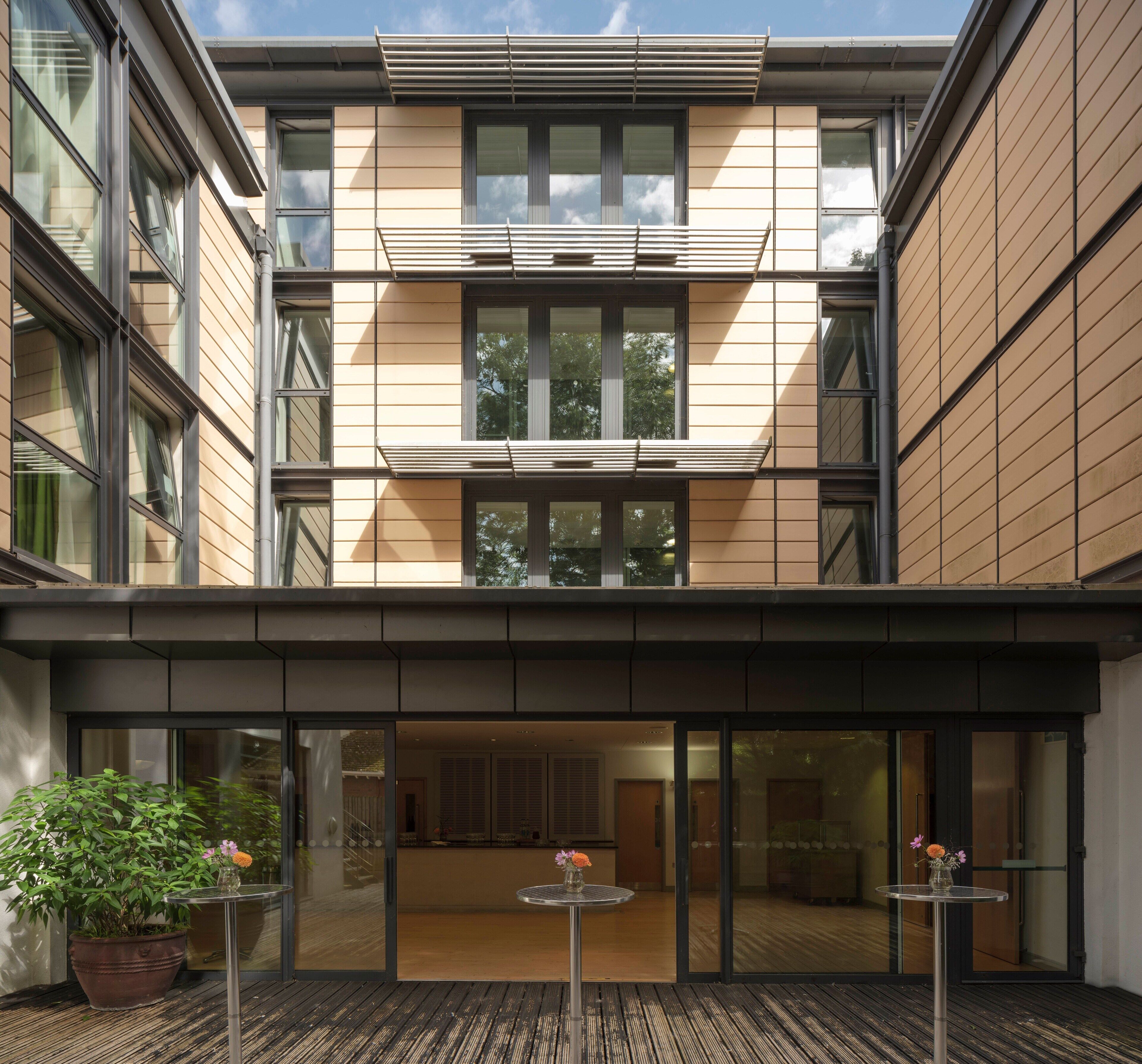 Ein modernes Gebäude mit großen Fenstern, einem überdachten Eingangsbereich und einer Holzterrasse mit Pflanzkübeln.