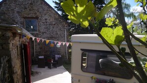 Un bâtiment en pierre doté d’un patio couvert, un camping-car et une petite table ornée d’un vase de fleurs.