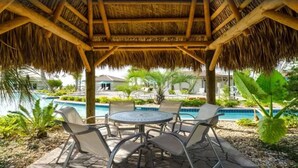 Een overdekt terras met tafel en stoelen, met uitzicht op een zwembad en een resort.