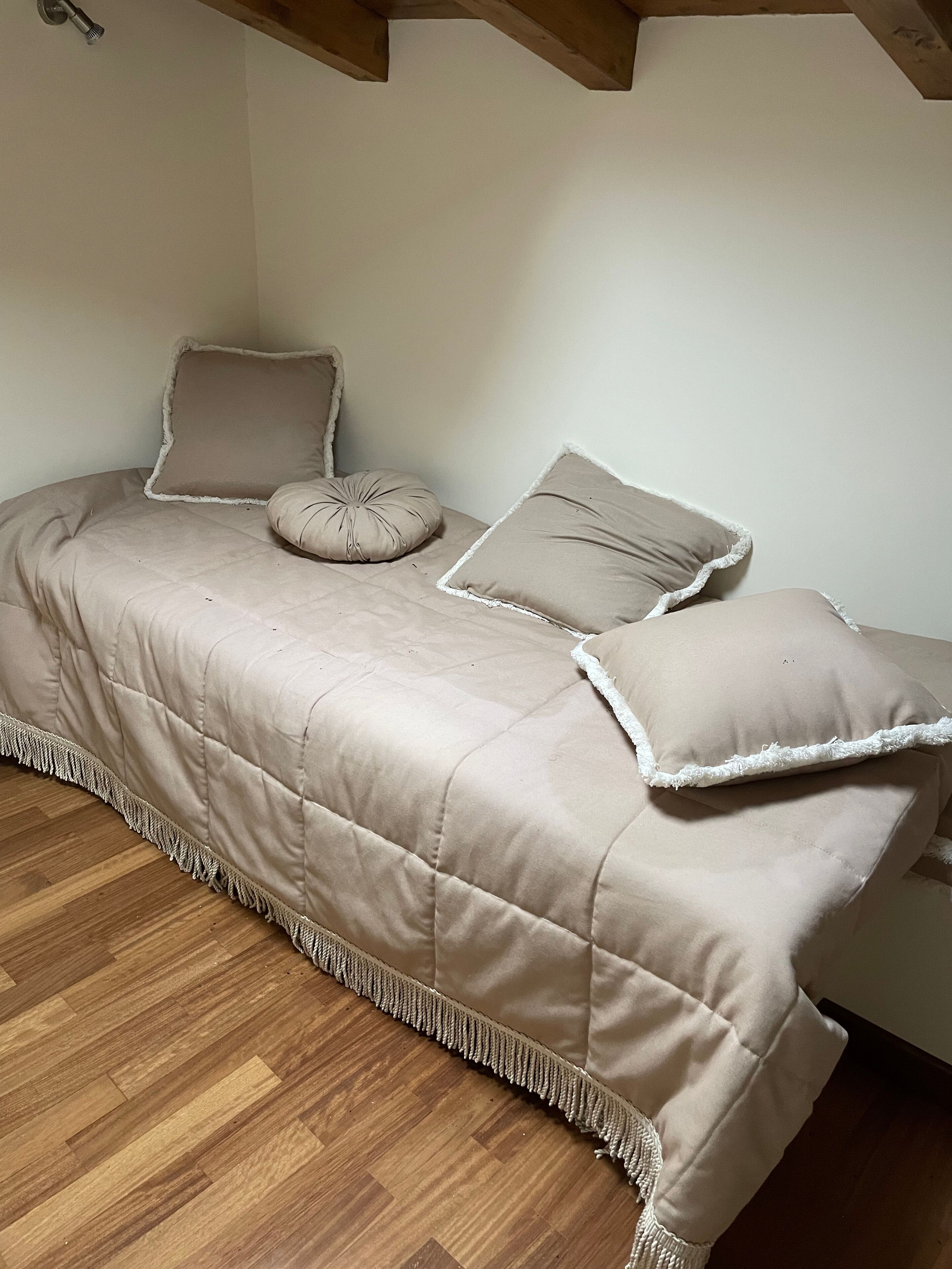 A small, cozy room with a bed featuring a quilted beige bedspread and white-framed pillows. The room has wooden flooring and a simple, clean design.