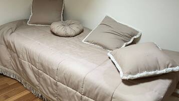 A small, cozy room with a bed featuring a quilted beige bedspread and white-framed pillows. The room has wooden flooring and a simple, clean design.