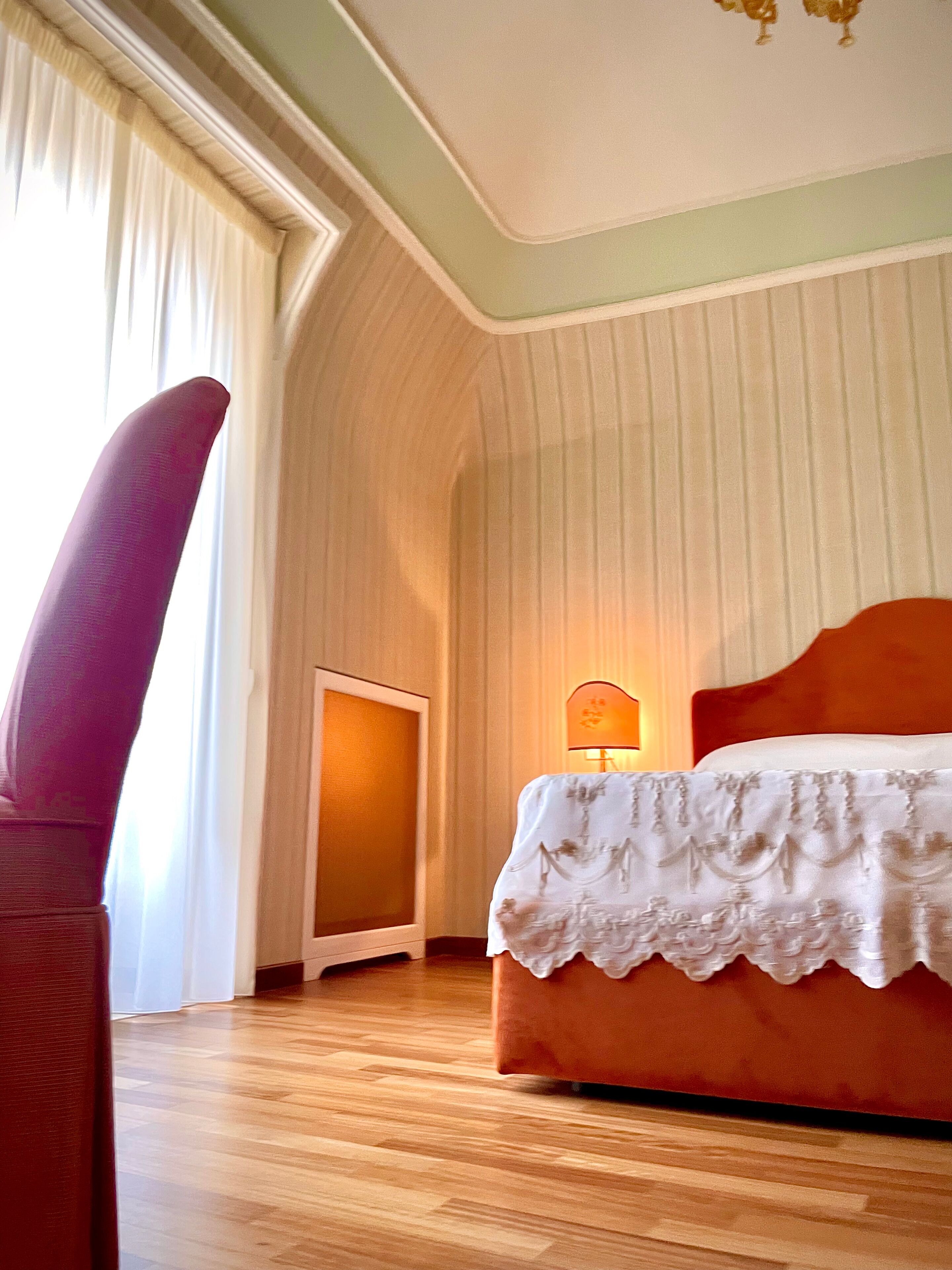 A bedroom with a wooden floor, a bed with a white bedspread, a nightstand with a lamp, and a window with curtains.