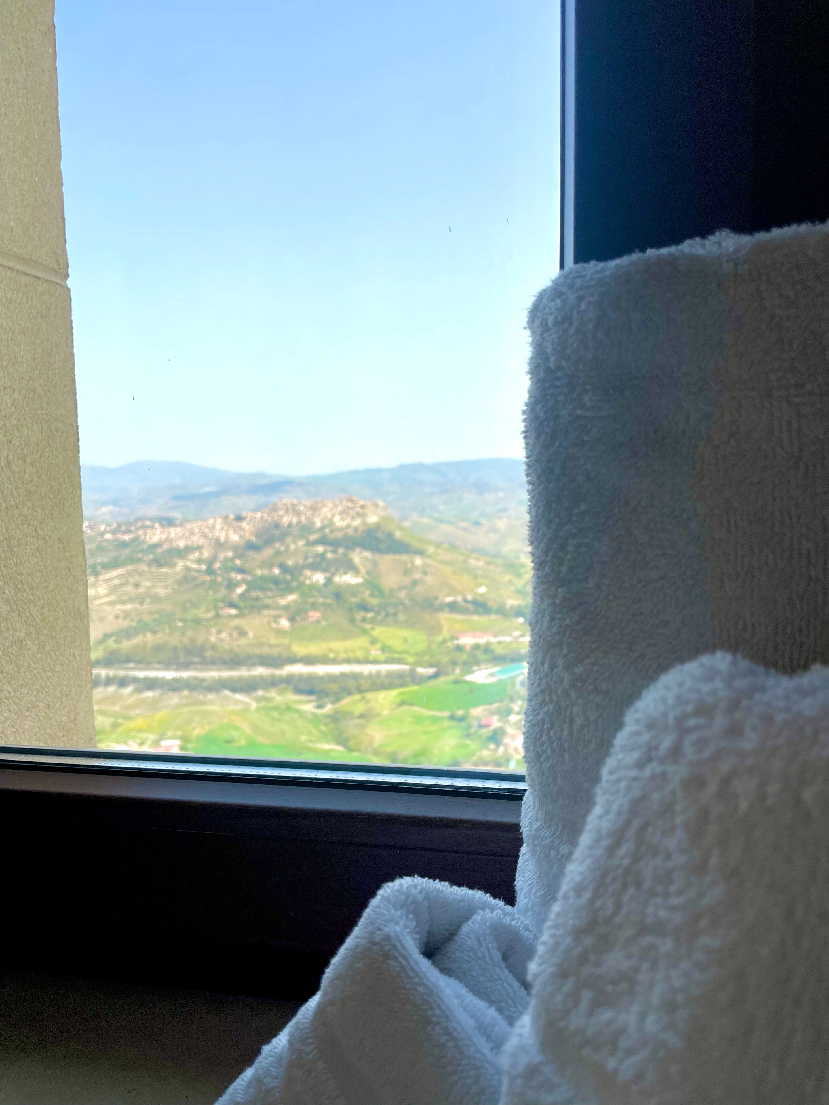 A window with a view of a green landscape and a white towel in the foreground.