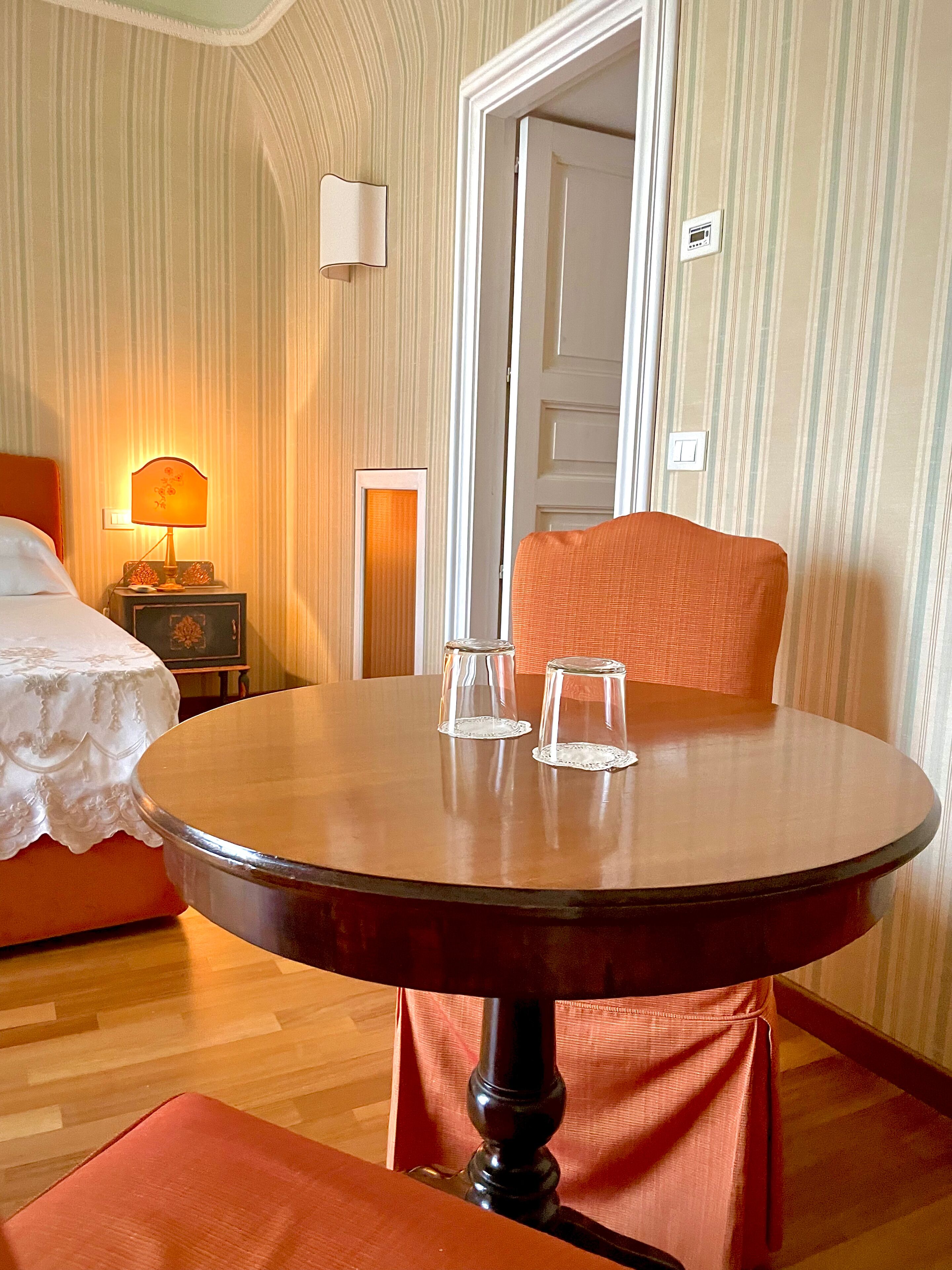 Habitación de hotel con una mesa redonda de madera, dos vasos transparentes, una silla tapizada en rojo, una cama con colcha blanca, una mesita de noche con lámpara y papel tapiz a rayas.