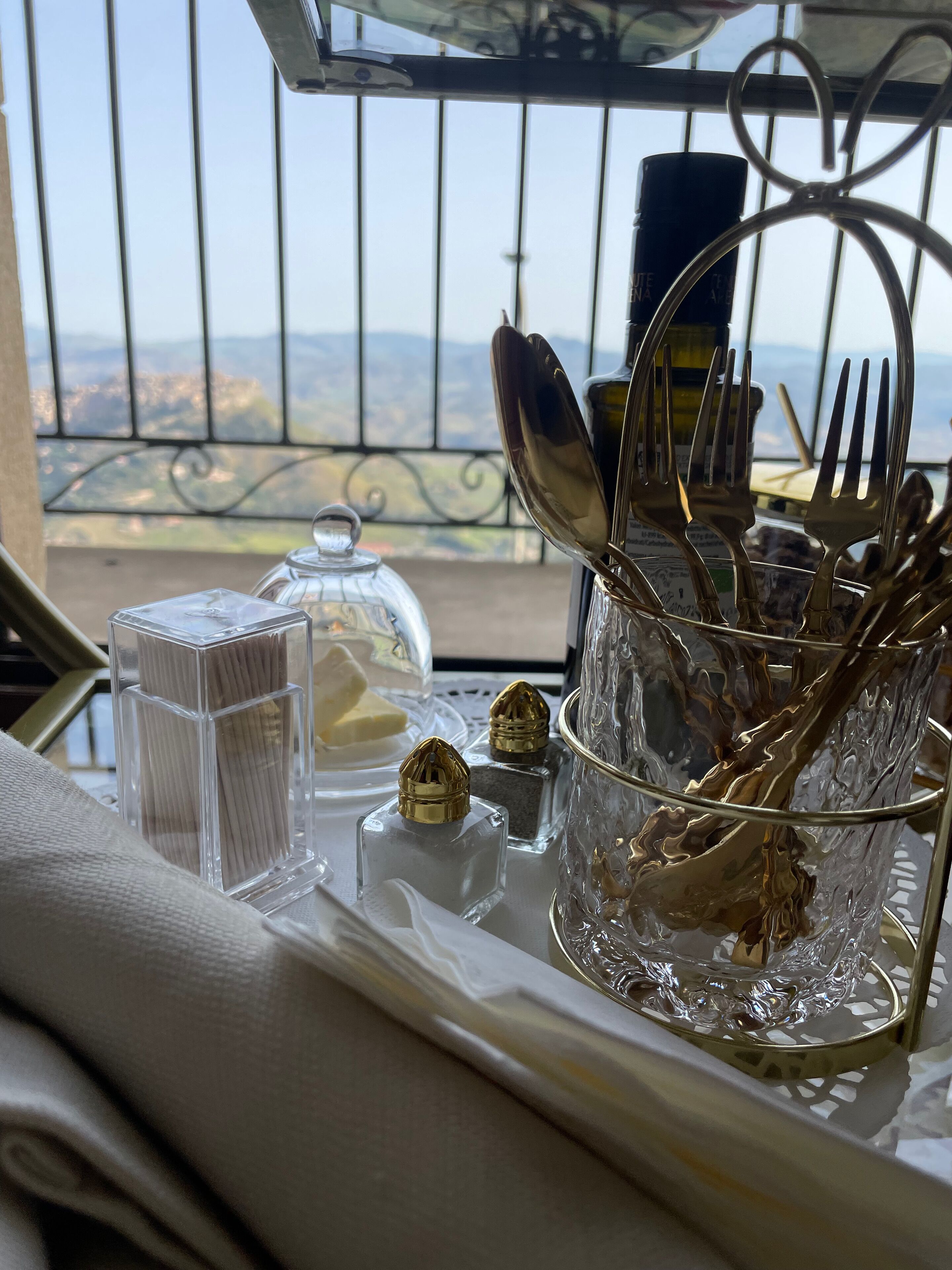 Una mesa de comedor con vistas a las montañas a través de una ventana, una bandeja metálica con cubiertos, saleros y pimenteros, y un recipiente de vidrio con una vela.