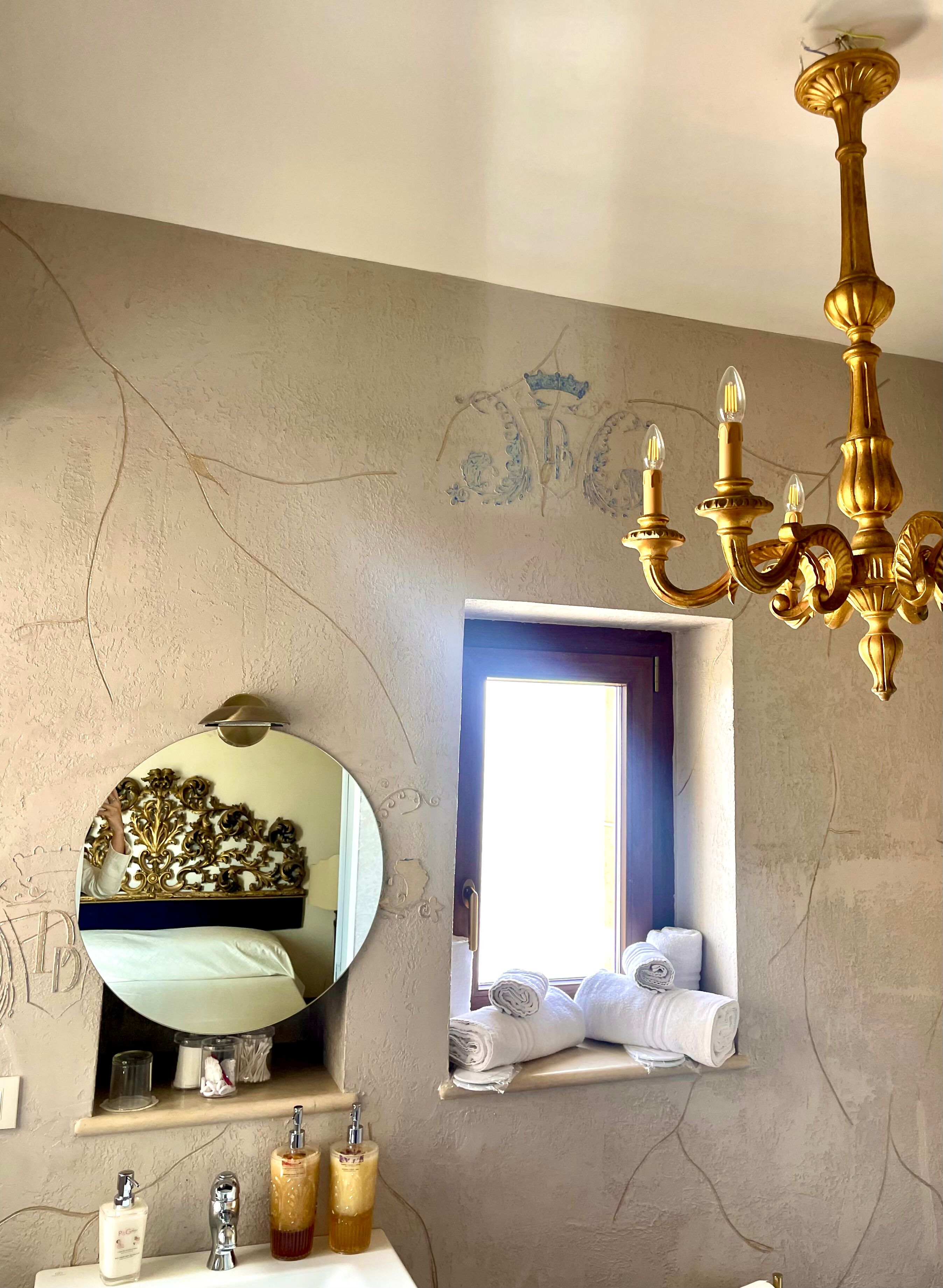 Un baño con un espejo redondo, una araña de cristal, una ventana con toallas y un estante con artículos de aseo.