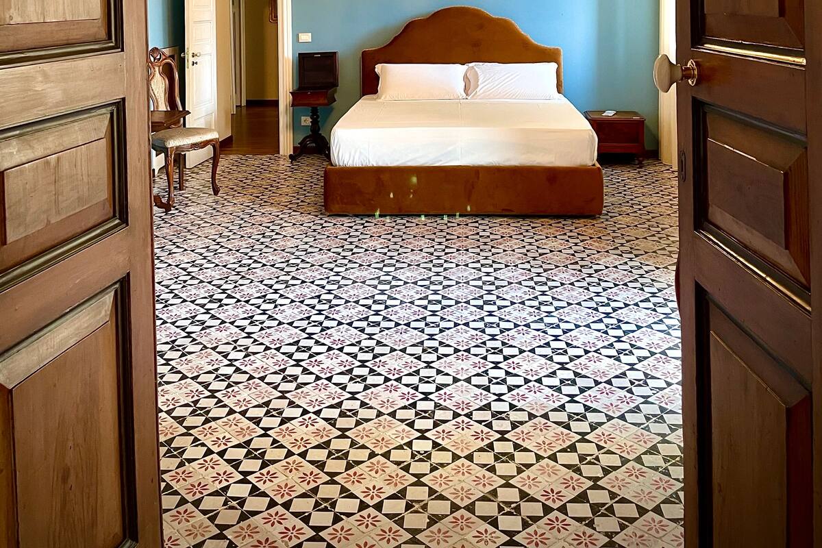 A bedroom with a large bed, a chandelier, a ceiling mural, and a patterned floor.