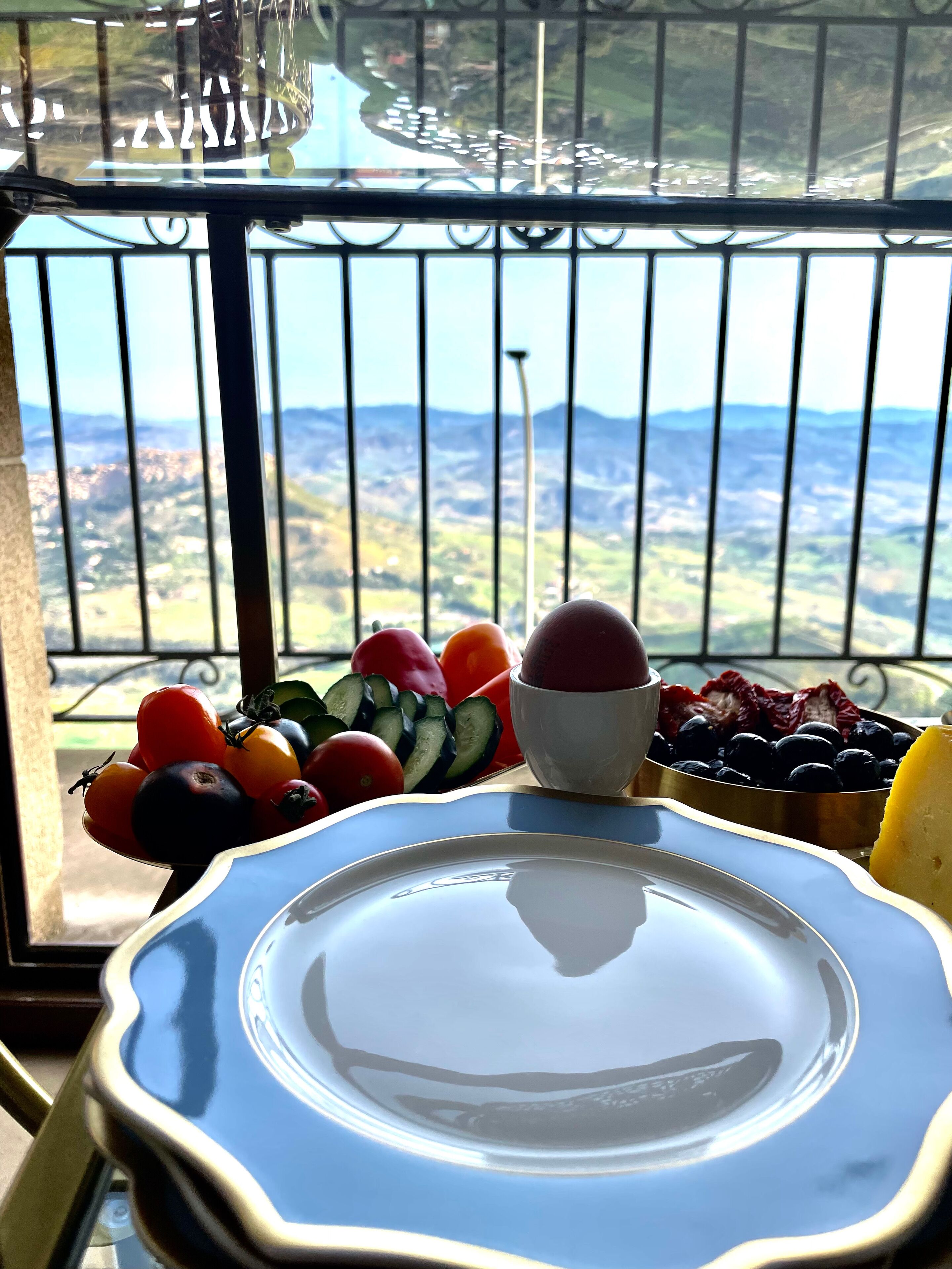 A balcony with a table set for a meal, featuring a plate, a bowl of fruit, and a bowl of grapes.