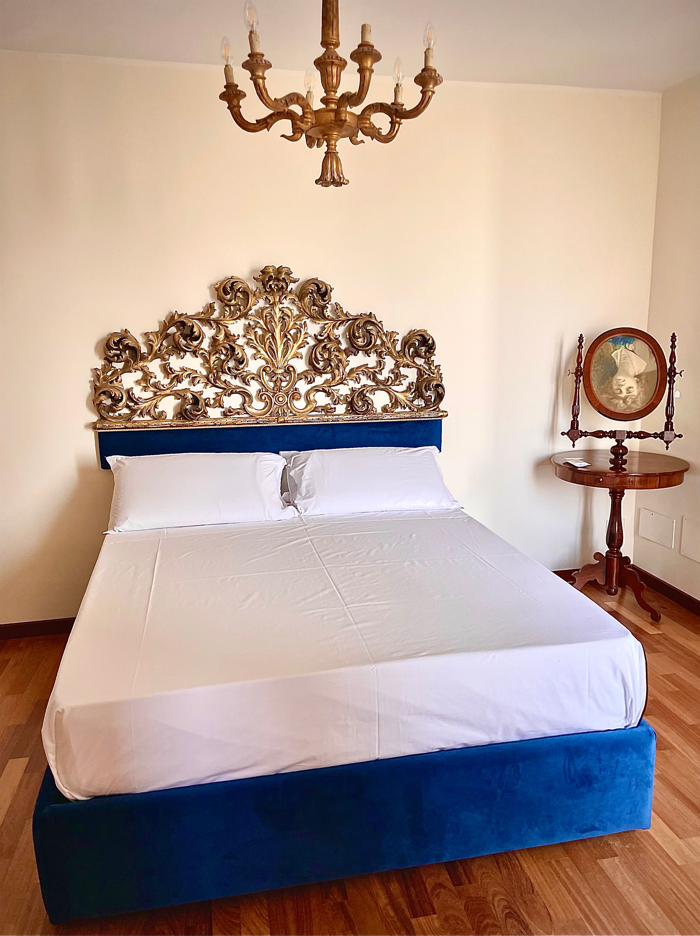 A bedroom with a large bed, a chandelier, a decorative headboard, and a round wooden table with a mirror.