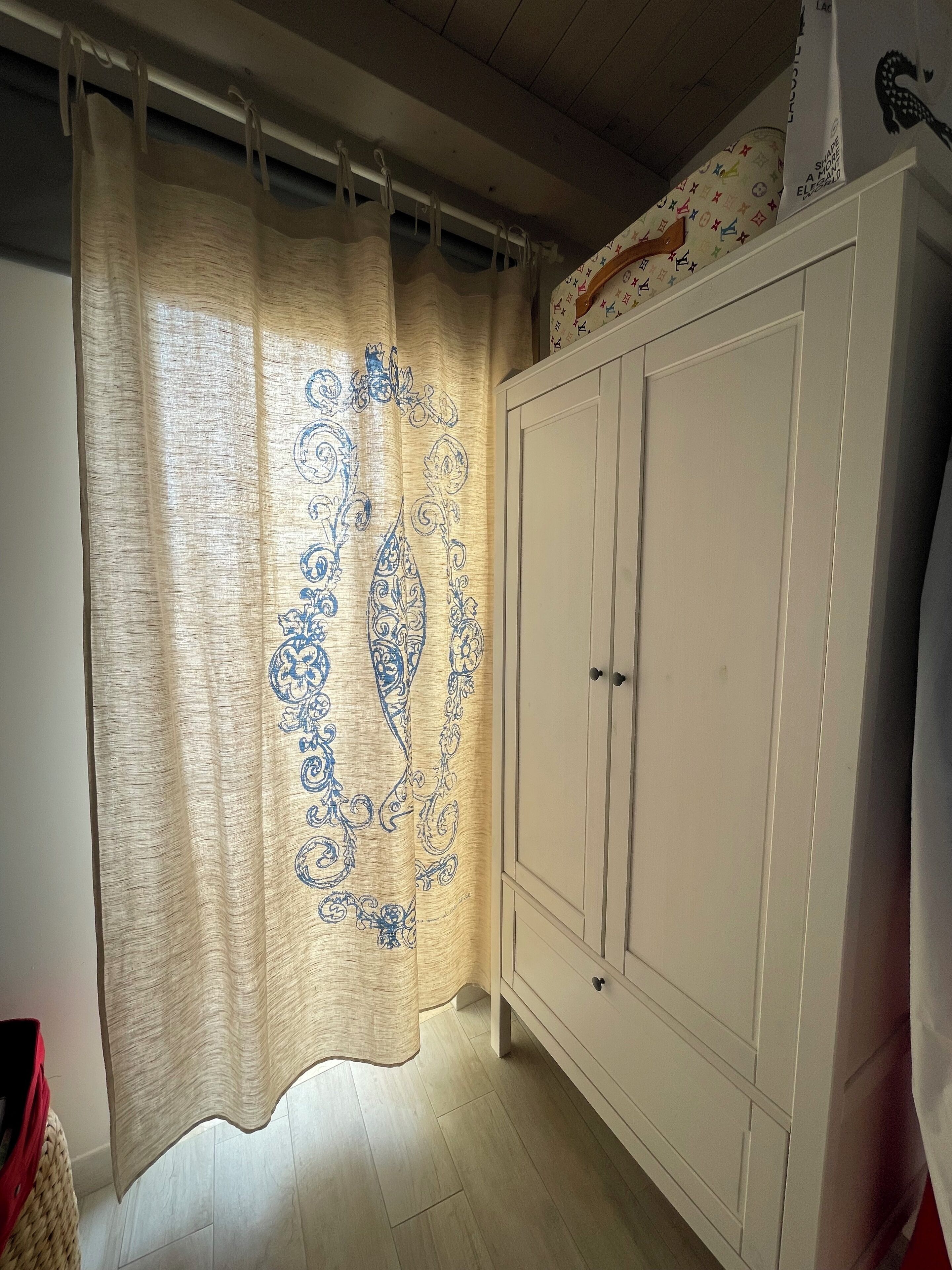 A room with a beige curtain featuring blue paisley patterns, a white wardrobe with two doors, and a tiled floor.