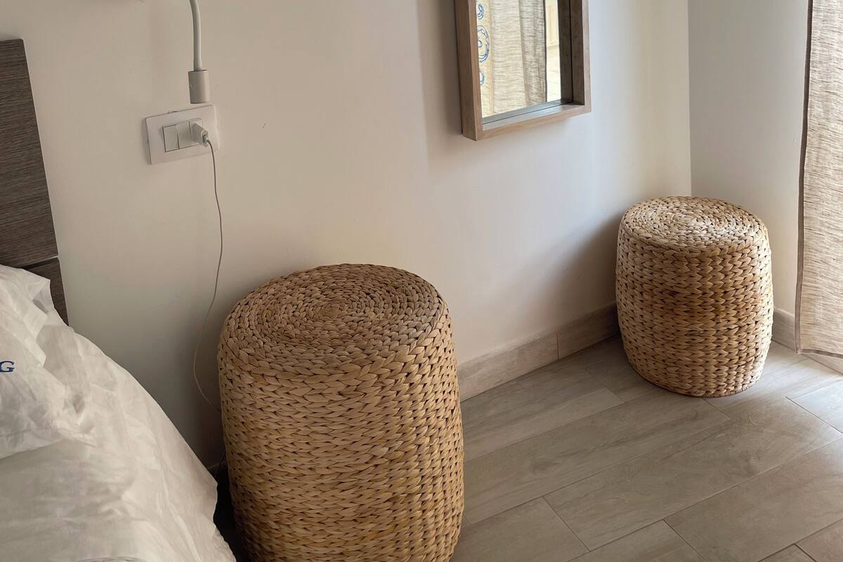 Une chambre avec un lit, un miroir fixé au mur, deux poufs en osier et une prise de courant.