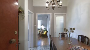 A dining area with a wooden table, chairs, and a chandelier. There is a door on the left and a hallway leading to another room. - Neasefessou25 (Kaisariani)