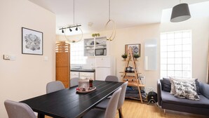 Een moderne woonkamer met een eettafel, grijze banken en een zwarte eettafel. Er zijn hanglampen, een ladder en een raam met jaloezieën.