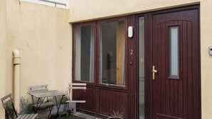 Een tweelaags huis met een donkerbruine voordeur, een kleine tuin met een groen gazon en een terras met een tafel en stoelen.