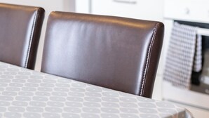 Un coin repas avec une table recouverte d’une nappe à motifs, deux chaises en cuir marron et, au fond, une cuisine dont le mur est agrémenté de carreaux rouges.