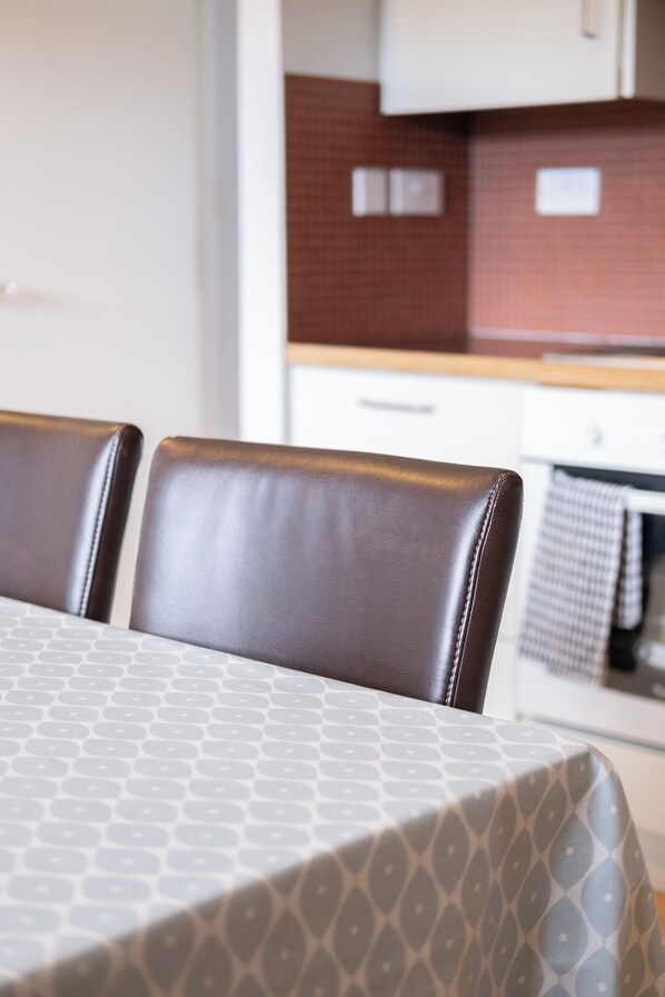 Een eethoek met een tafel bedekt met een geruit tafelkleed, twee bruine leren stoelen en een keuken op de achtergrond met een rode betegelde spatwand.