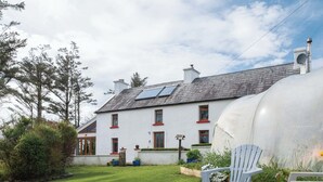 Uma casa branca com telhado inclinado e painéis solares, um reservatório de água grande e um gramado com três galinhas.
