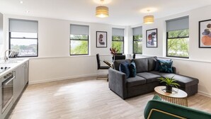 Ein modernes Wohnzimmer mit grauem Sofa, einem grünen Sessel und einem runden Couchtisch. Zwei Pendelleuchten, ein Essbereich mit schwarzem Stuhl und eine Küche mit weißer Arbeitsplatte und Spüle.