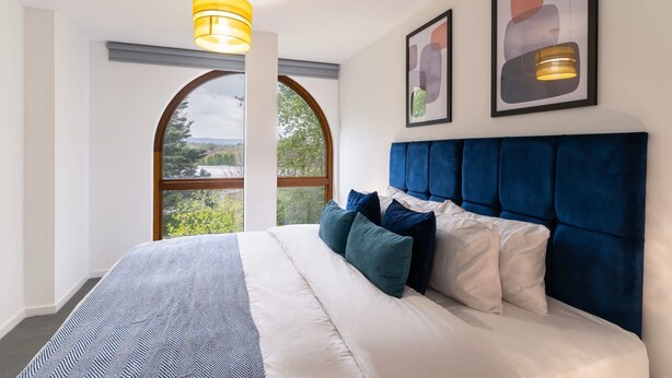 Ein Schlafzimmer mit einem großen Bett, einem blauen Kopfteil und Blick durch ein Fenster.