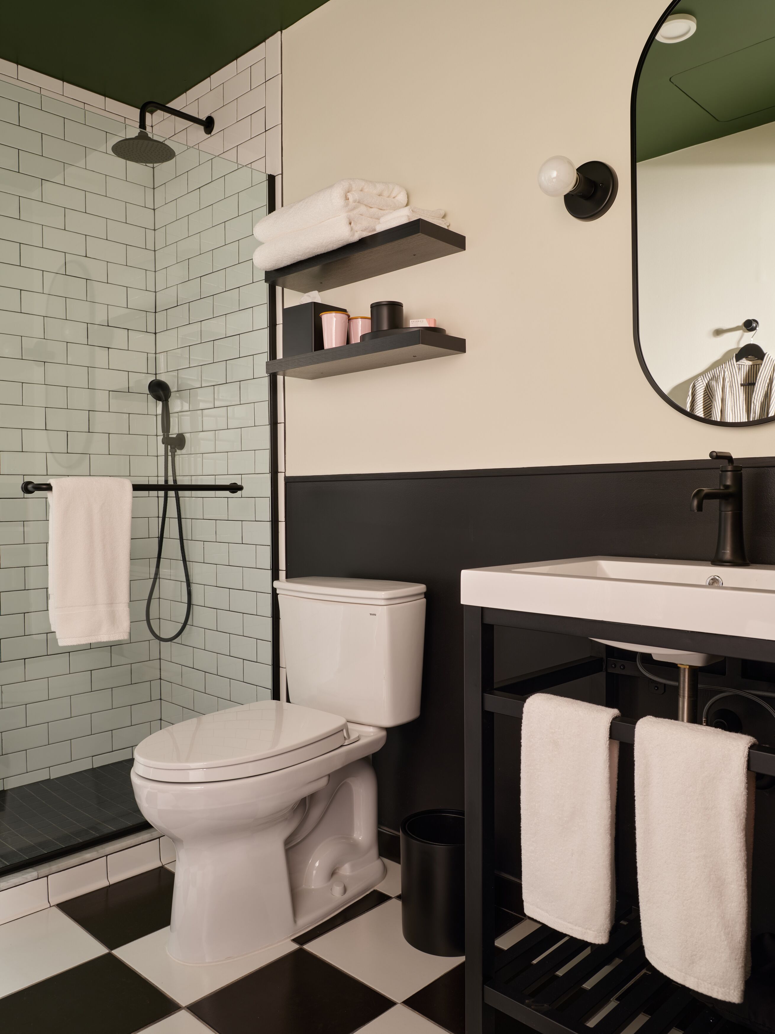 Un baño moderno con inodoro blanco, lavamanos negro y ducha. Toallas blancas cuelgan de un perchero y hay un estante con artículos de aseo.